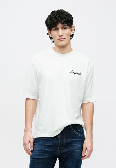 Balta kokvilnas T-krekls ar īsām piedurknēm, uz augšējās kreisās puses ir melns "Dsquared2" logotips. Modelis valkā tumši zilas džinsa bikses.