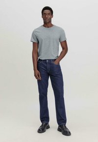Grijze katoenen t-shirt met korte mouwen, gecombineerd met donkerblauwe denim jeans. Model staat rechtop met handen in de zakken, en draagt zwarte schoenen.