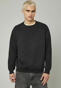 Sweat-shirt noir à col rond, fabriqué dans un material doux, avec des poignets et un ourlet côtelés, porté sur une chemise crème, avec une coupe décontractée.
