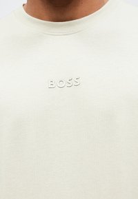 Šviesiai smėlio spalvos sportinis megztinis su ribotu, apvaliu kaklu ir iškiliniu, įspaustu „BOSS“ logotipu ant krūtinės. Minkšta tekstūra ir laisvas modelis.