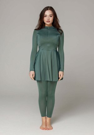 Woman with long brown hair stands barefoot wearing a fitted, long-sleeve green dress with matching leggings against a plain gray background.