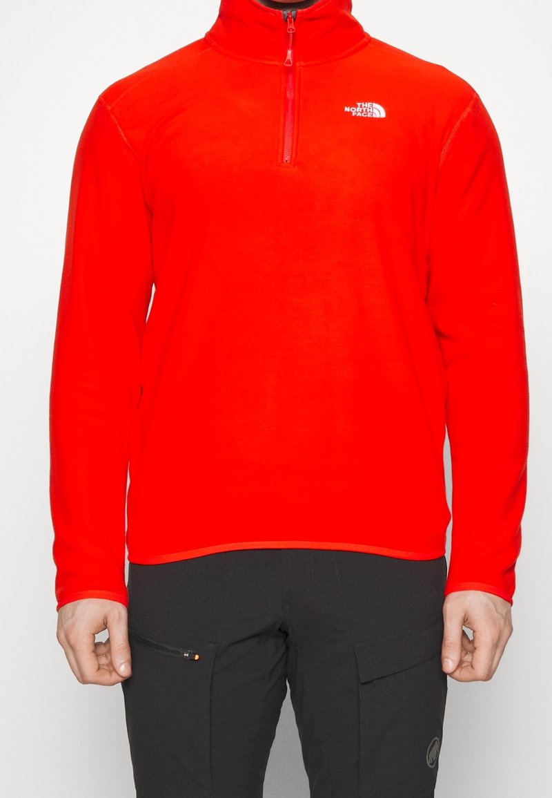 Personne portant un fleece rouge vif à fermeture quart de zip avec le logo The North Face et un pantalon noir avec poches zippées et cargos.