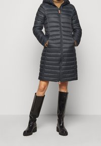 Mørkegrå dunjakke med hætte, med quiltet tekstur og lynlås foran. Kombineret med knæhøje sorte støvler og en tanfarvet sweater.