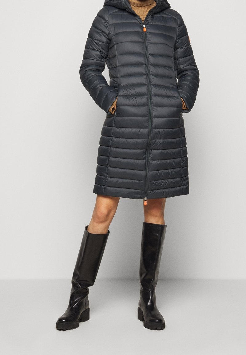 Mørkegrå dunjakke med hætte, med quiltet tekstur og lynlås foran. Kombineret med knæhøje sorte støvler og en tanfarvet sweater.