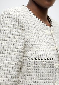 Veste en tweed blanc avec un motif texturé, dotée d'un bord noir le long de l'encolure et d'une petite poche avec des détails de couture noire.