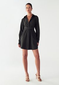 Tumedast rohelisest kangast särgikleit, millel on krae, pikad varrukad, vöökoht ja ees taskud, koos heledate beezide kontsakingadega.