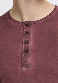 Burgunderfarbenes Henley-Shirt mit rundem Ausschnitt, vier braunen Knöpfen an der Knopfleiste und einem weichen, strukturierten Stoff.