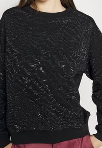 Sudadera negra con un patrón metálico texturizado, cuello redondo y puños acanalados. El diseño cuenta con un brillo sutil sobre una tela suave.