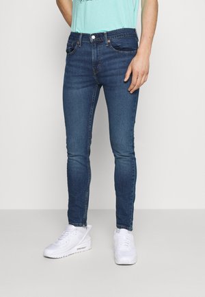 Homme portant un jean slim bleu foncé et des baskets blanches, debout devant un fond clair uni.
