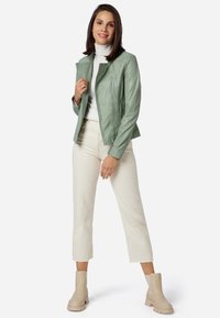 Mintgrön läderjacka med dragkedja, sidofickor och strukturerad passform, bärs över en vit polotröja och krämfärgade korta byxor.