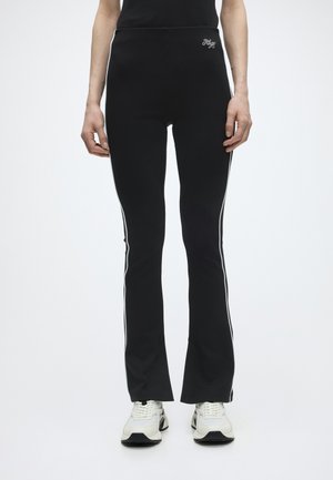 Persona che indossa pantaloni neri aderenti con strisce bianche laterali e scarpe da ginnastica bianche, in piedi davanti a uno sfondo chiaro e uniforme.