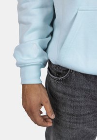 Felpa azzurra con maniche a costine, caratterizzata da un piccolo logo a forma di diamante bianco sulla manica, abbinata a jeans grigi scuri con texture.