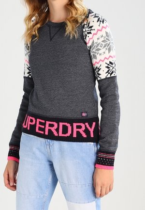 Kobieta w ciemnoszarym swetrze z wzorzystymi białymi i różowymi rękawami oraz jasnoróżowym napisem "SUPERDRY" na dolnym brzegu, w połączeniu z jasnoniebieskimi dżinsami.