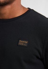 Zwarte geribbelde sweatshirt met een vierkant label met "Petrol Ind" in oranje, geplaatst op een textuurstof. Ronde halslijn en een aansluitend ontwerp.