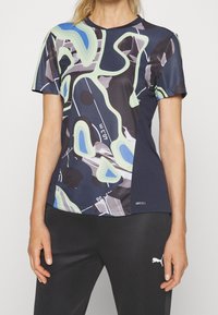 Vrouw die een sportshirt met korte mouwen draagt met abstracte blauwe, groene, zwarte en witte patronen en zwarte Puma-broek.