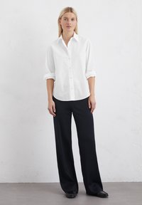 Camicia bianca con maniche arrotolate, abbinata a pantaloni neri a gamba larga. Tessuto liscio, colletto classico e taglio dritto.