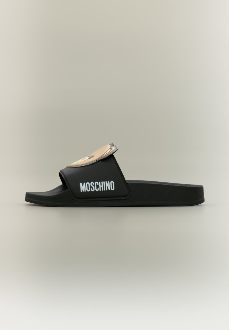 Sandale slide noire Moschino avec détail d'ours en dessin animé beige sur la bride et logo blanc sur le côté, vue de profil sur un fond neutre.