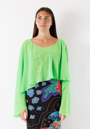 Donna dai lunghi capelli castani indossa una maglietta ampia di colore verde brillante e una gonna colorata con fiori blu e forme astratte rosse, viola e verdi.