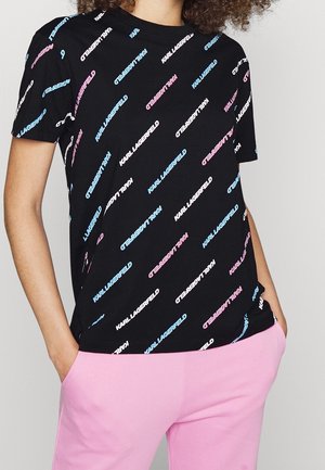 Persona que lleva una camiseta negra con texto diagonal repetido "KARL LAGERFELD" en azul, rosa y blanco, combinada con pantalones rosas, manos en los bolsillos.