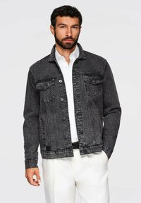 Giacca in denim nera con due tasche sul petto, chiusura a bottoni e un finish leggermente sbiadito. Il modello indossa una camicia bianca sotto.