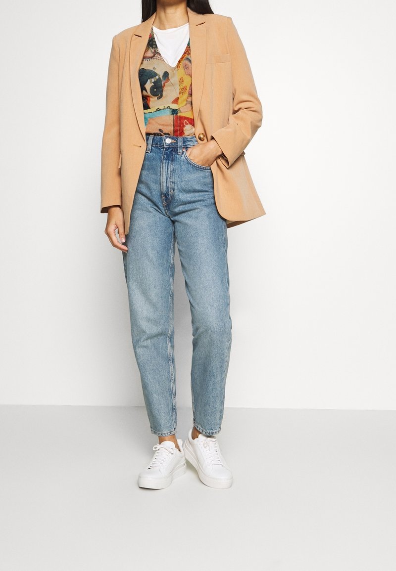 Femme portant un blazer beige sur un top multicolore à motifs, un jean taille haute bleu clair et des baskets blanches, debout avec une main dans la poche.
