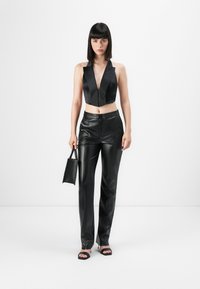 Top halter em satin preto, cropped, com decote profundo, combinado com calças de pele pretas de cintura alta e sapatos de salto pretos com tiras, segurando uma pequena bolsa.