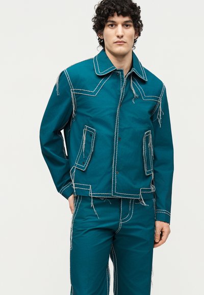 Άνδρας που φοράει τζιντζάκετ και παντελόνι σε πετρόλ απόχρωση με υπερμεγέθη άσπρα ράμματα και φθαρμένες άκρες, που στέκεται μπροστά σε απλό ανοιχτό φόντο.