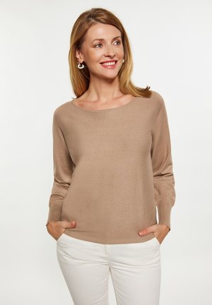 Lichtbruine trui, zachte textuur, brede halslijn, verfittende mouwen, gedragen met witte broek. Model staat met handen in de zakken en glimlacht.