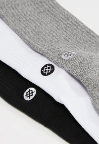 Three pairs of ankle socks displayed, in gray, white, and black. Each has a small, round embroidered logo on the side. Textured material.