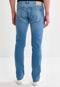 Hombre con jeans ajustados de color azul claro y zapatillas blancas, de pie mirando hacia atrás contra un fondo blanco liso.