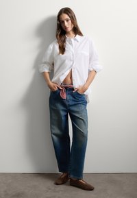 Camisa blanca de botones con mangas remangadas, jeans de mezclilla azul con una corbata roja estampada en la cintura y zapatos planos marrones. Fondo neutro.