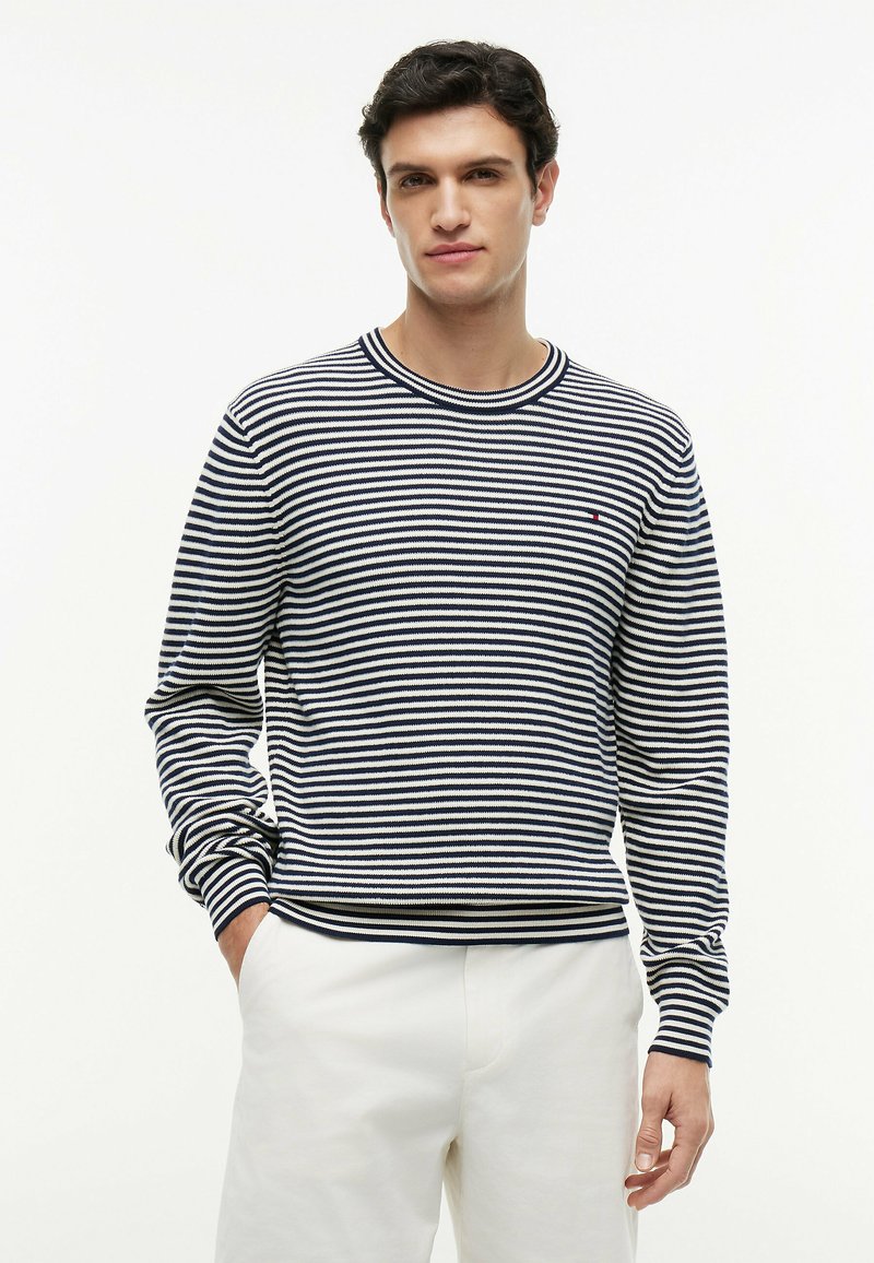 Homme portant un pull à manches longues rayé bleu marine et blanc et un pantalon blanc, debout avec une main dans la poche sur un fond uni.