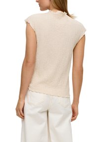 Beige, gestructureerde top met korte mouwen en een gerimpelde kraag, gecombineerd met witte wijde broekspijpen. De stof heeft een lichte wafelstructuur.
