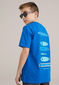 Kék póló, hátul grafika található, amely különböző kék árnyalatú surfboardokat ábrázol. Pamut anyag, rövid ujjú, klasszikus szabású.
