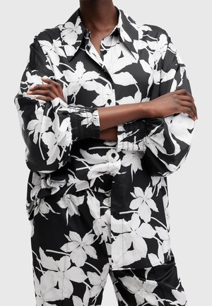 Persona con una camisa de manga larga de estampado floral en blanco y negro y pantalones a juego, con los brazos cruzados sobre el pecho.