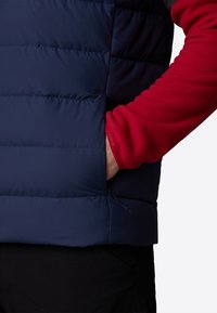 Puffet marineblå jakke med en rød fleecemanchet. Har en lynlåslomme og elastisk kantbånd til justering. Glat tekstur med quiltet design.