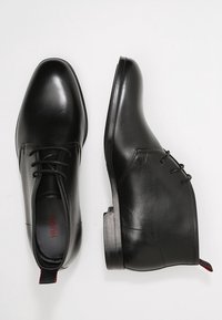 Černé kožené šněrovací boty s hladkým povrchem, kulatou špičkou a nízkým, vrstveným podpatkem. Obsahují jemný červený akcent na patním poutku.