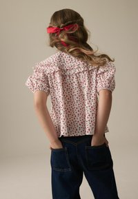 Weißes Blusenshirt mit rotem Blumenmuster, gerafftem Kragen und kurzen Puffärmeln, kombiniert mit dunklen Jeans. Haare mit einem roten Band zusammengebunden.