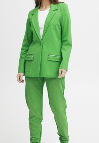 Grön kavaj och matchande byxor i ett mjukt material. Kavajen har en enkel knäppning och två framsidan dragkedjefickor.