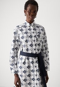 Abito camicia bianco con motivo geometrico blu navy, maniche lunghe, colletto e cintura coordinata. Presenta tasche e chiusura frontale con bottoni.
