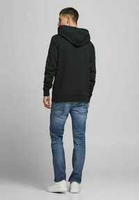 Sudadera negra con capucha y un corte holgado, combinada con jeans de mezclilla azules ajustados y zapatillas blancas, vista desde atrás.