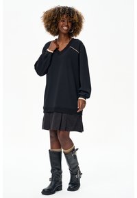 Uśmiechnięta kobieta o kręconych włosach nosi luźny czarny sweter z dekoltem w serek, plisowaną spódnicę, beżowe skarpetki oraz czarne kozaki sięgające połowy łydki z klamrami.