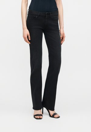 Γυναίκα που φοράει στενά μαύρα τζιν και μαύρα ψηλοτάκουνα σανδάλια με ανοιχτά δάχτυλα, στέκεται μπροστά σε απλό ανοιχτόχρωμο φόντο.