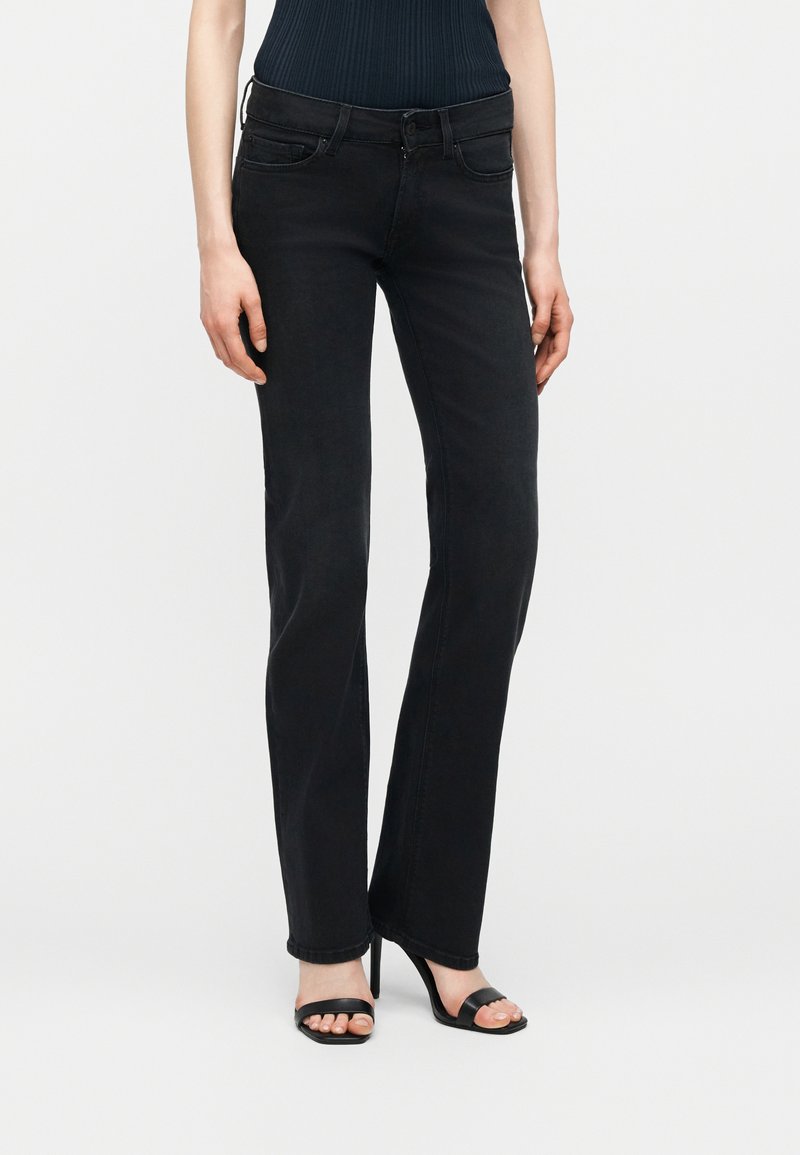 Γυναίκα που φοράει στενά μαύρα τζιν και μαύρα ψηλοτάκουνα σανδάλια με ανοιχτά δάχτυλα, στέκεται μπροστά σε απλό ανοιχτόχρωμο φόντο.