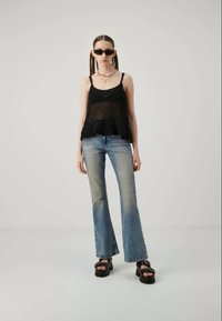 Μαύρο πλεκτό τοπ με φρου-φρου λεπτομέρεια στο τελείωμα, συνδυασμένο με ανοιχτό μπλε καμπάνα τζιν. Το μοντέλο φοράει χοντρά μαύρα σανδάλια και σκούρα γυαλιά ηλίου.