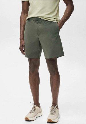 Homme portant un short vert olive, des baskets beiges et une chemise claire, debout avec une main dans la poche devant un fond uni.