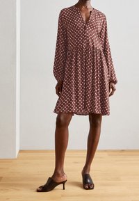 Vestido marrón con lunares, de manga larga, escote en V y cintura fruncida. Acompañado de mules negros abiertos en la punta. Fondo de suelo de madera.