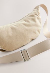 Bēšs korduroja vidukļa soma ar gludu tekstūru, aprīkota ar krēmkrāsas regulējamu siksnu un metāla sprādzi. Kompakts un izliekts dizains.