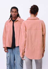Roze corduroy overhemd met een knoopsluiting aan de voorkant, twee borstzakken en lange mouwen. Toont zowel voor- als achteraanzichten. Zachte textuur met verticale strepen.