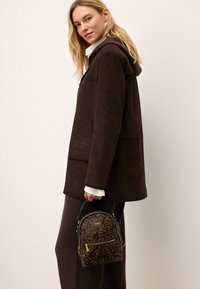 Kohvatooni nubukist takjak, millel on kapuuts, ülepaisutatud sobivus ja kaks esikotti. Hoides käes väikest musta ja pruuni leopardimustriga seljakotti kullast aktsentidega.